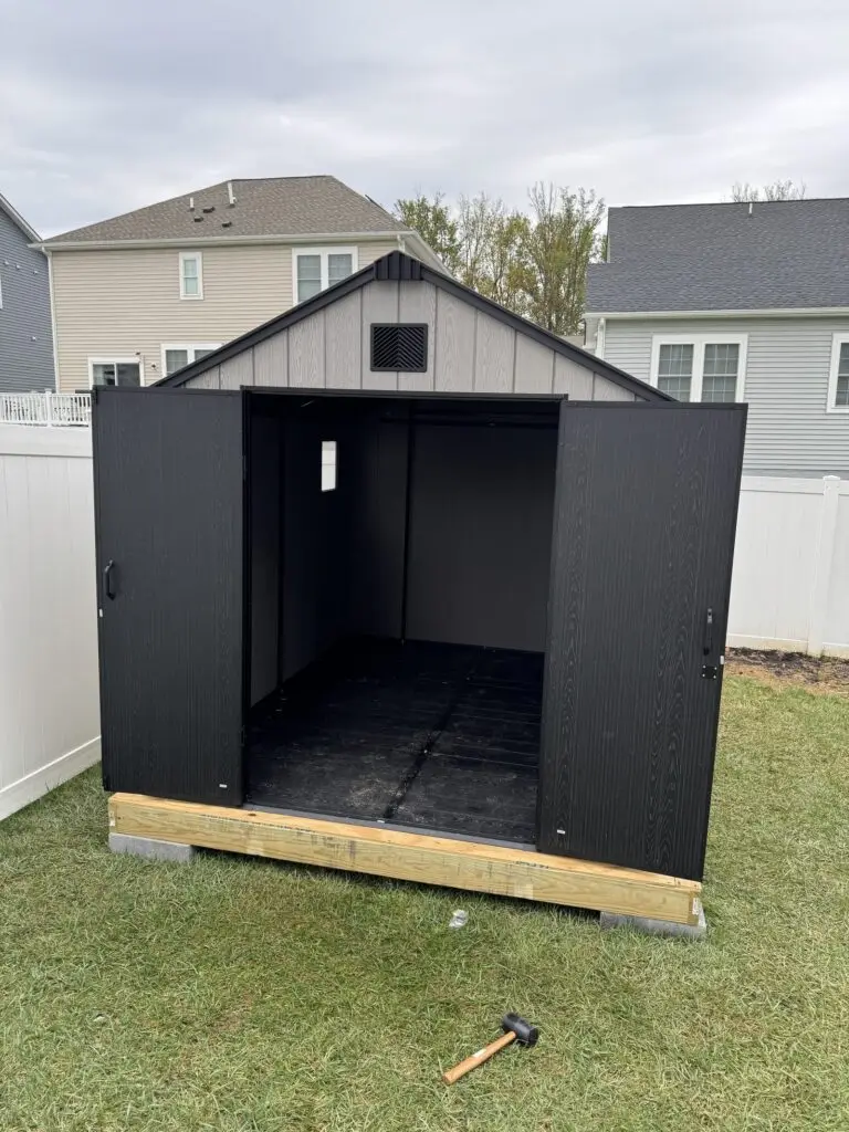 Black and gray outdoor storage shed assembled on a wood base in a fenced backyard